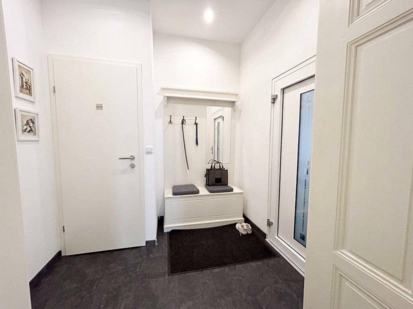 Heller Eingangsbereich mit Garderobe, Spiegel, Sitzbank und dunklem Fliesenboden.