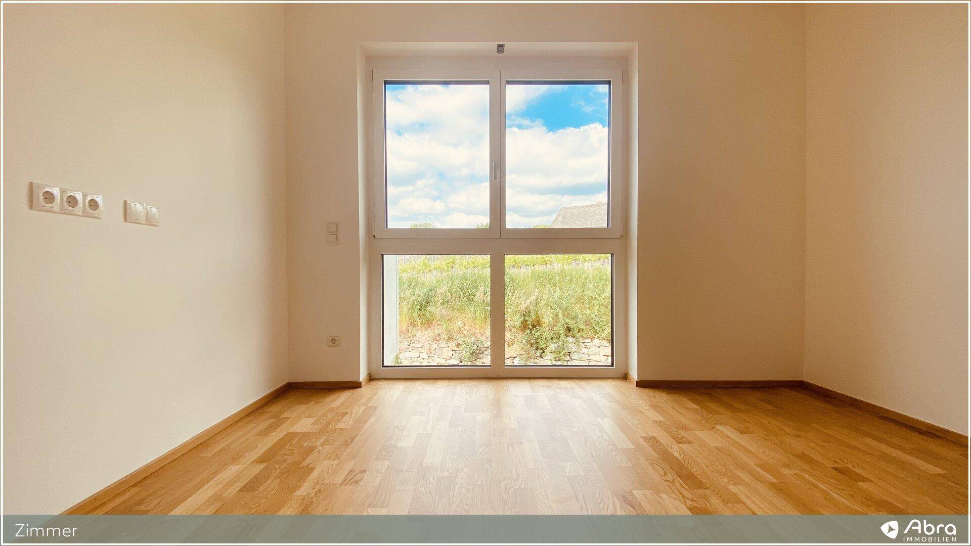 Helles Zimmer mit Holzboden und großem Fenster, das einen Blick ins Grüne bietet.