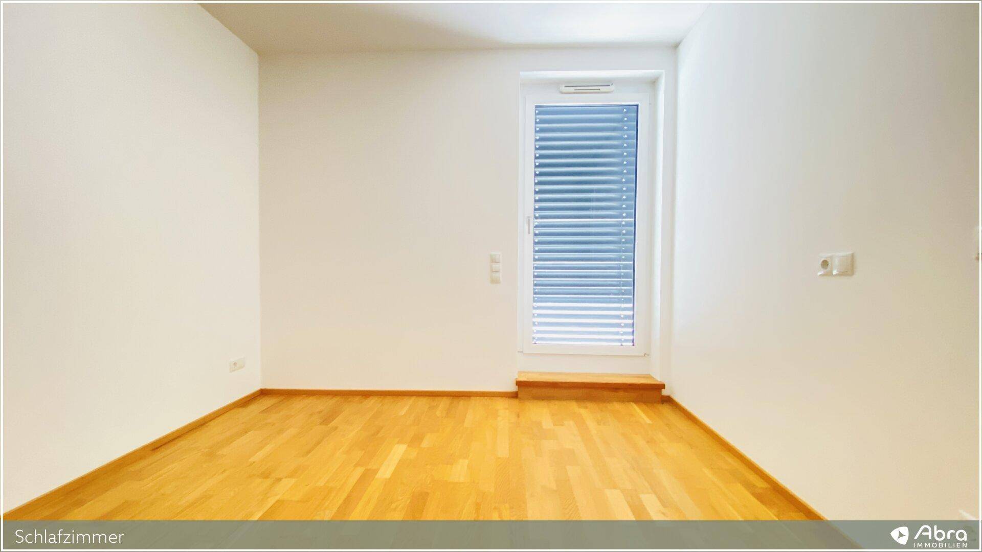 Leeres Schlafzimmer mit hellem Holzboden und einem Fenster mit Jalousien.