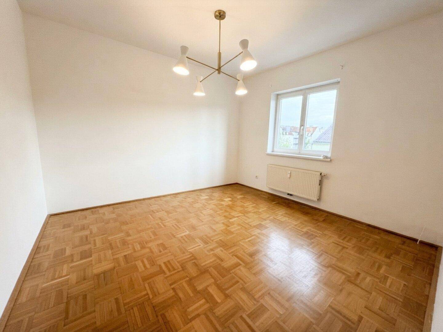 Heller Raum mit Parkettboden, großem Fenster und moderner Deckenleuchte, ideal als Wohnzimmer.
