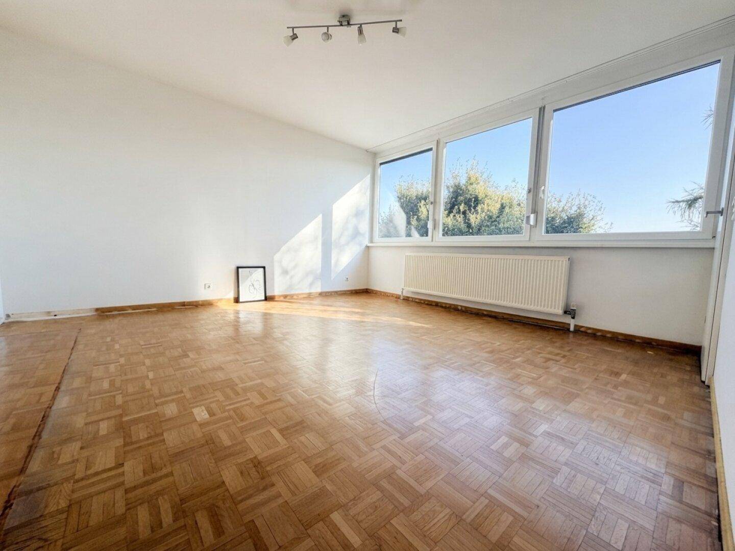 Geräumiges Wohnzimmer mit Fischgrätparkett, großen Fenstern und viel Sonnenlicht, bereit zur Einrichtung.