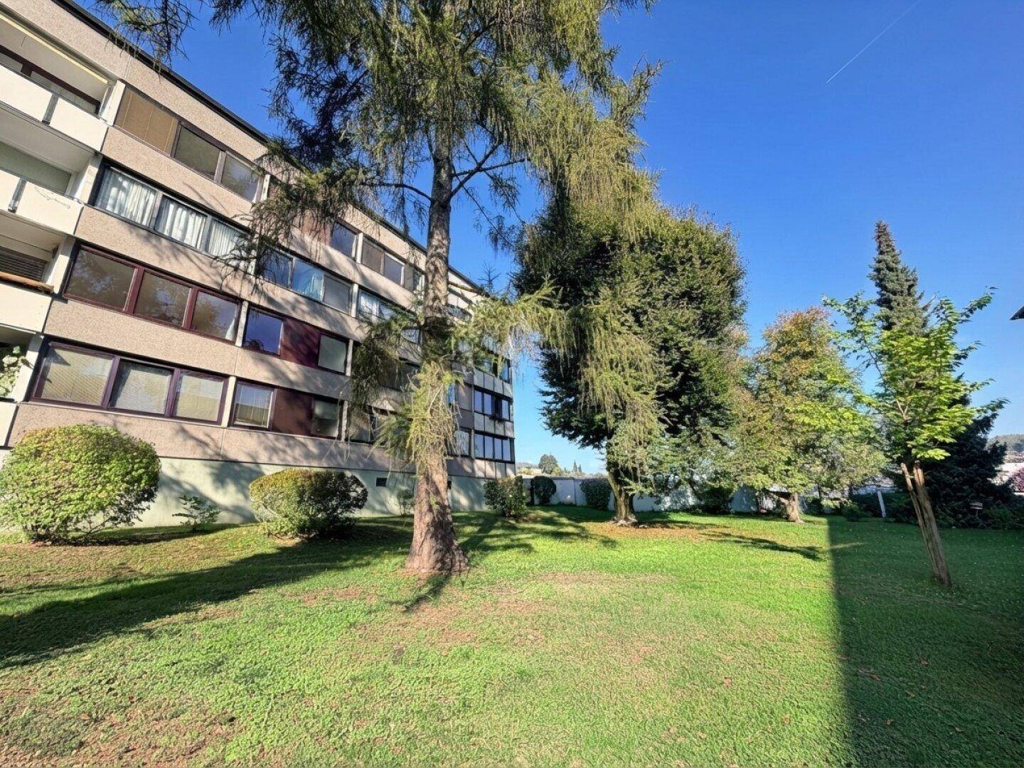 Mehrstöckiges Wohngebäude mit gepflegter Rasenfläche und Bäumen im Vordergrund unter blauem Himmel.