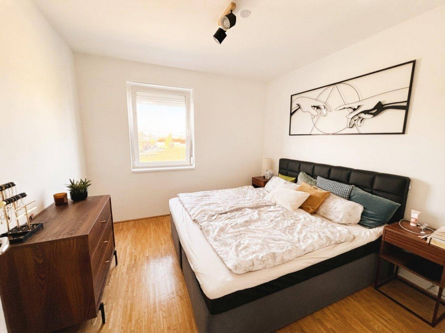 Helles Schlafzimmer mit Doppelbett, Holzkommode und großem Fenster für viel Tageslicht.