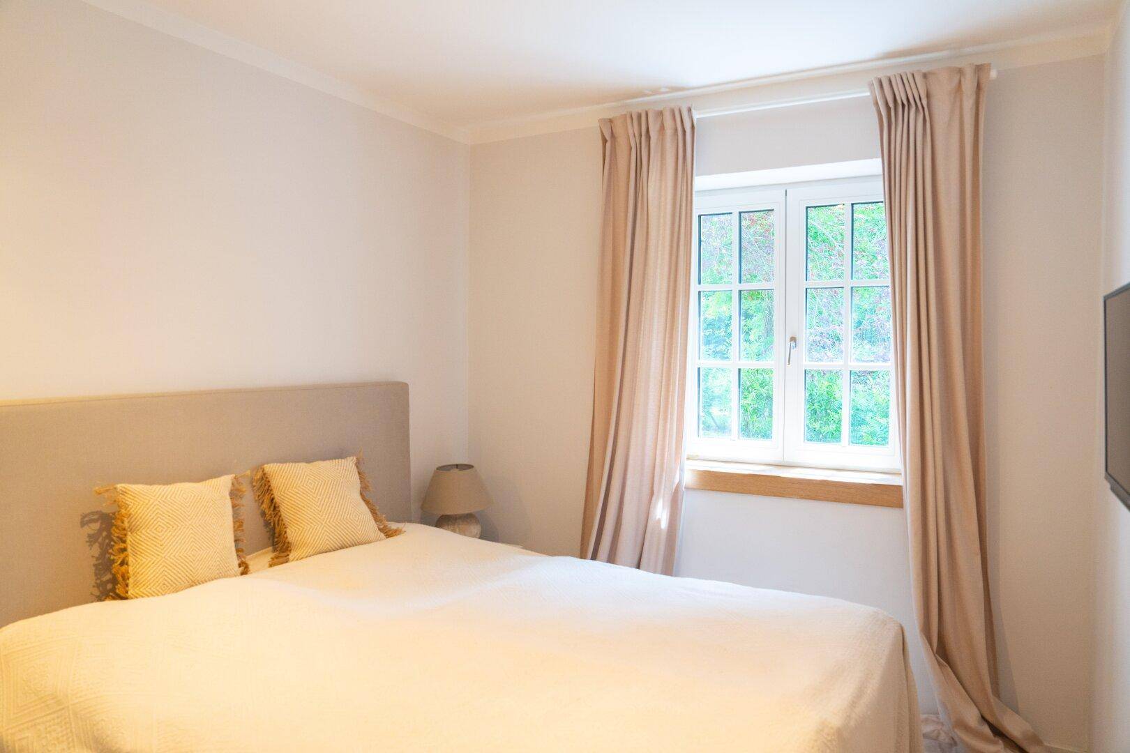 Helles Schlafzimmer mit Doppelbett, Vorhängen und Fenster mit Blick ins Grüne.