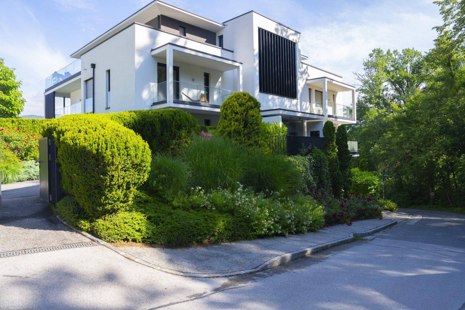 Moderne weiße Wohnanlage mit mehreren Balkonen und gepflegter Gartenlandschaft an einer ruhigen Straße.