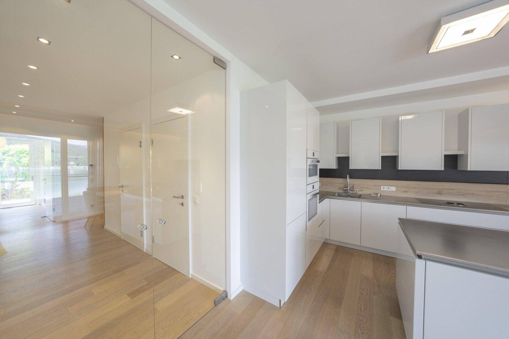 Moderne weiße Küche mit glänzenden Fronten und Holzboden, angrenzend an einen hellen Flurbereich.