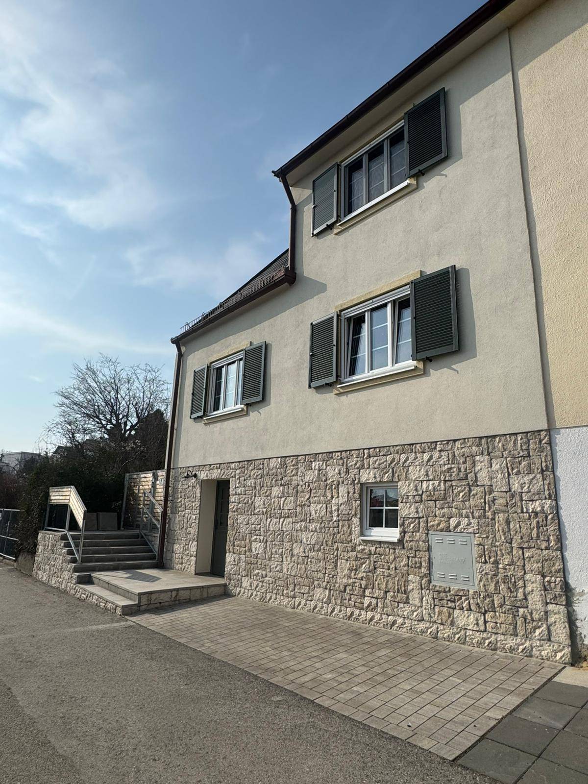 Seitenansicht des Hauses mit Steinsockel, Fensterläden und gepflastertem Eingangsbereich.