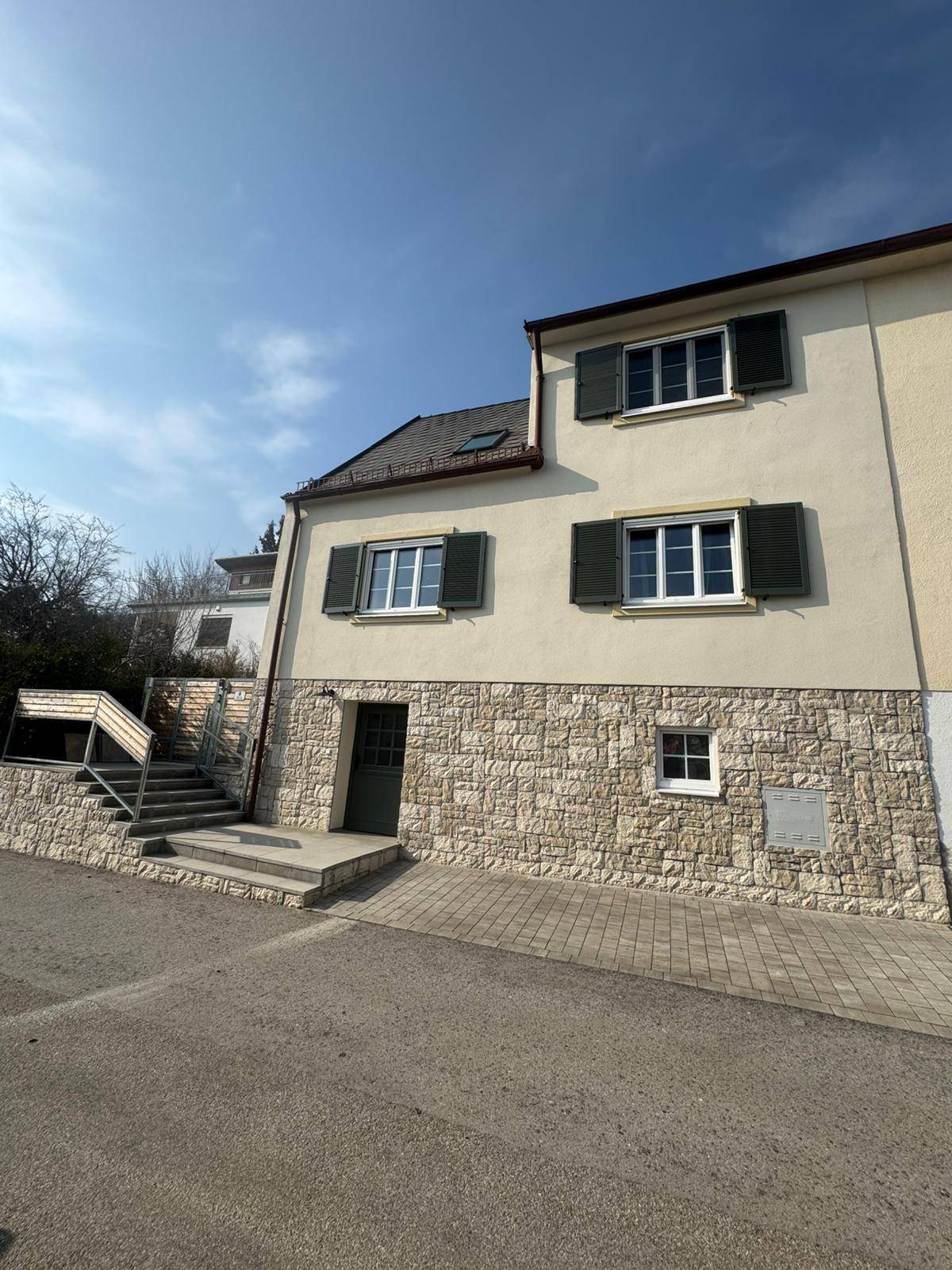 Ansicht der Hausfassade mit Steinsockel, Fensterläden und gepflastertem Zugangsbereich.