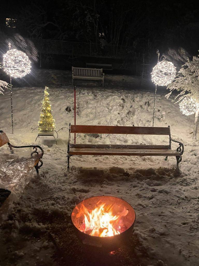 Winterlicher Garten bei Nacht mit Feuerstelle, beleuchteten Kugeln und verschneiten Bänken.