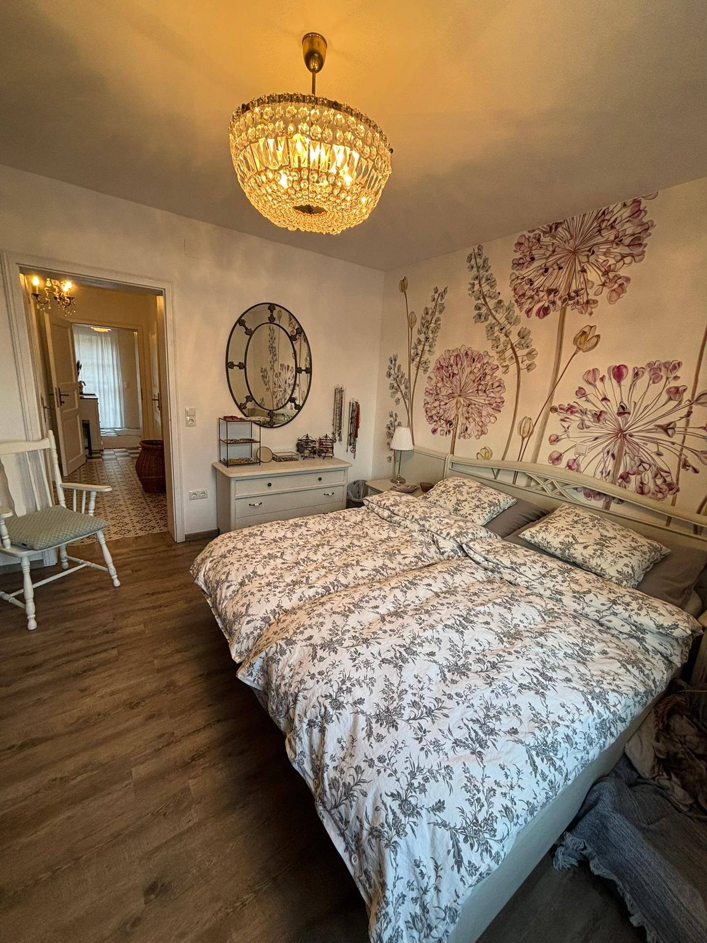 Schlafzimmer mit floraler Wanddekoration, Doppelbett und Kronleuchter, mit Blick in den Flur.