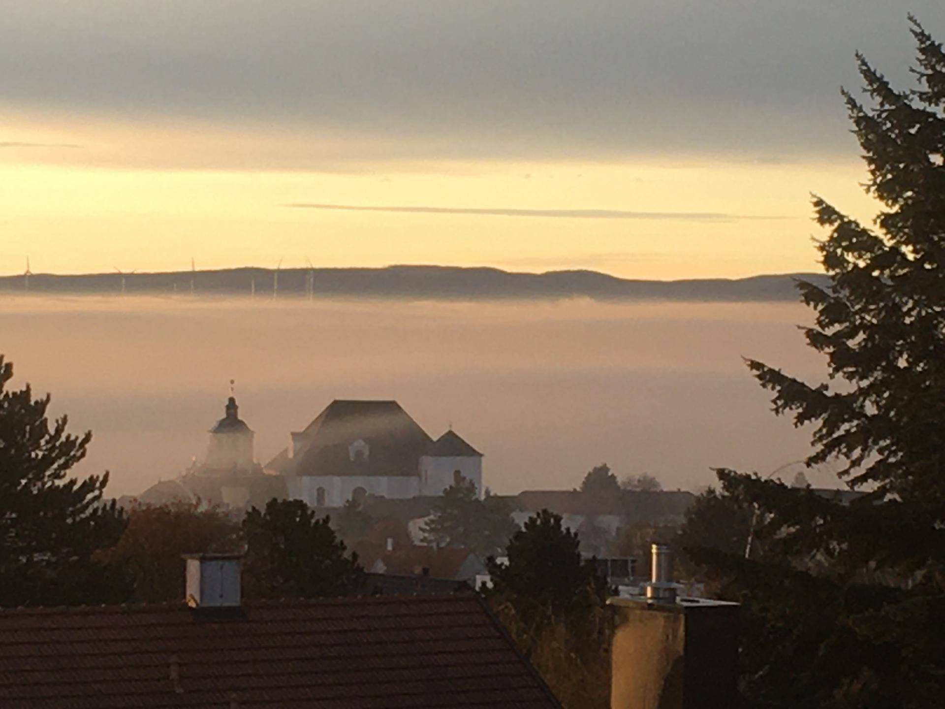 Malerischer Ausblick über eine nebelverhangene Landschaft mit Kirche und Windrädern am Horizont.