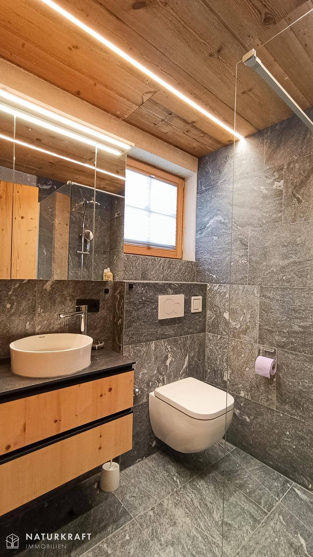 Modernes Badezimmer mit dunklen Fliesen, Waschtisch, wandhängendem WC und Fenster.