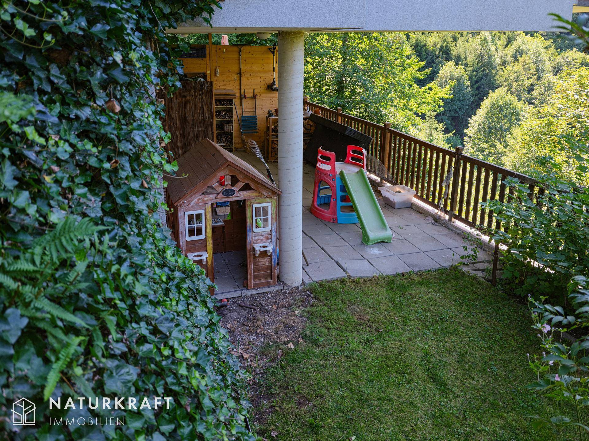 Gartenbereich mit Spielhaus und Rutsche, umgeben von Bäumen und üppigem Grün.