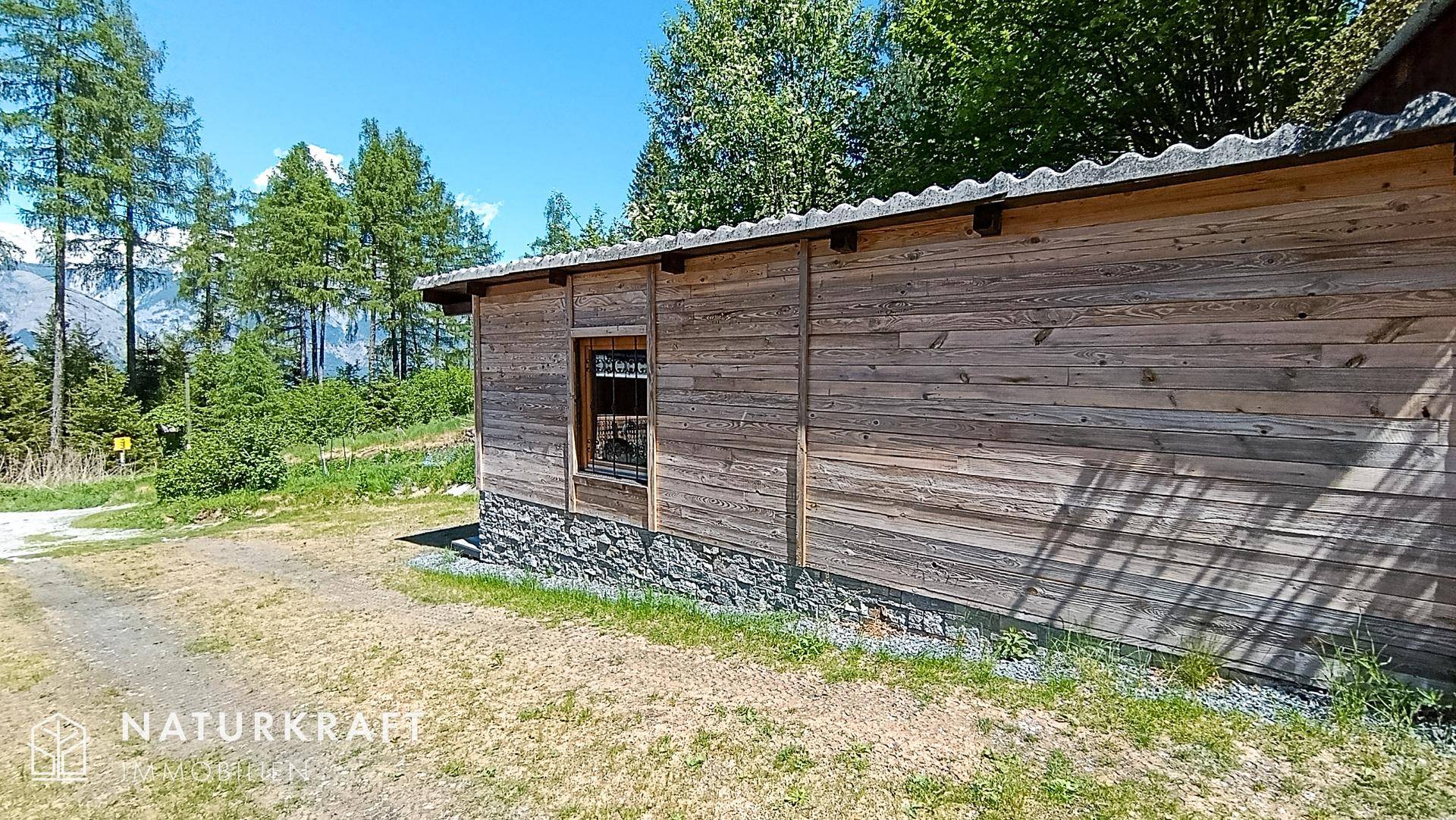 Seitenansicht eines Holzgebäudes mit Steinsockel und grüner Umgebung unter strahlendem Himmel.