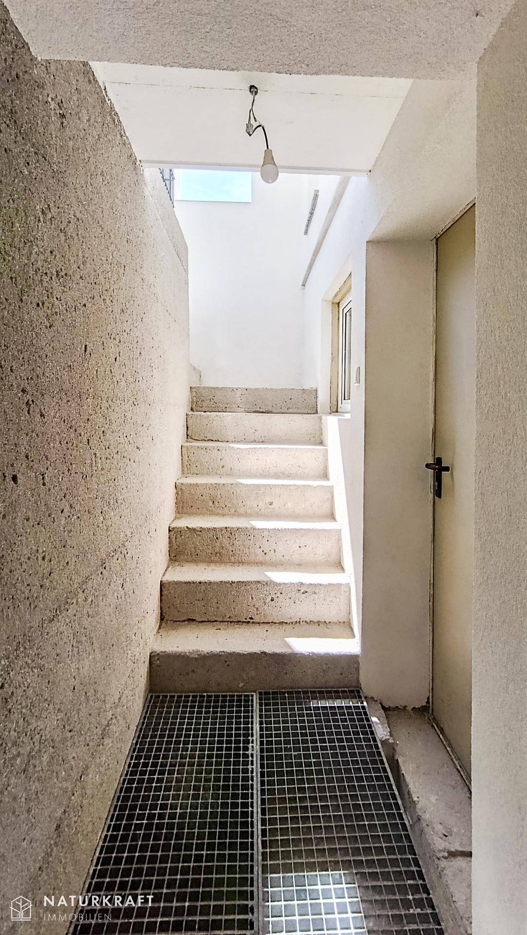 Betontreppe führt hinunter in einen hellen Kellerbereich mit Gitterrostboden.
