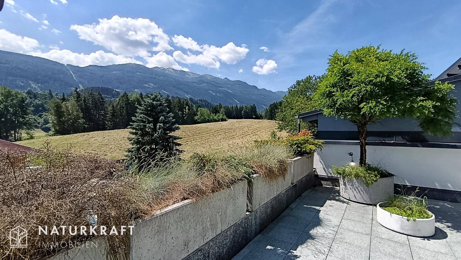Weitläufiger Bergblick von einer Terrasse mit Bepflanzung und weiten Feldern im Hintergrund.