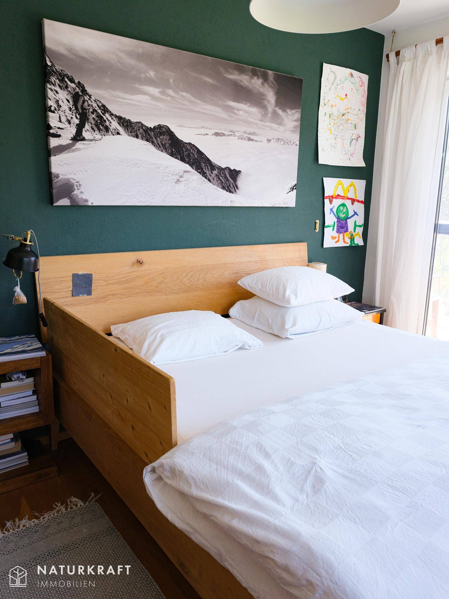 Schlafzimmer mit Holzbett, grüner Akzentwand und großem Wandbild, das eine Berglandschaft zeigt.