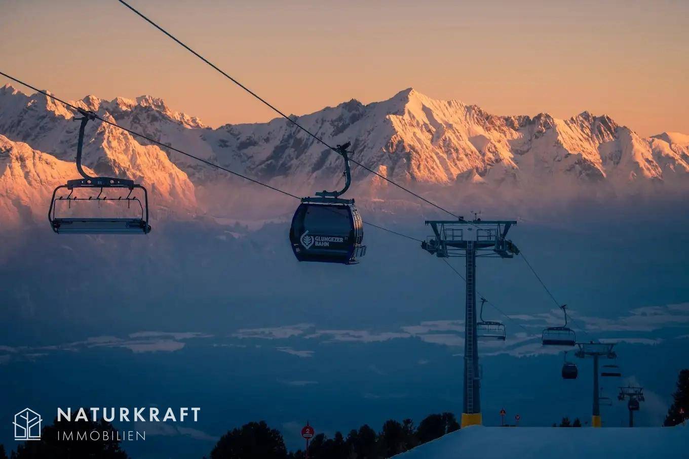 Skilift und Gondel vor einer beeindruckenden Bergkulisse im warmen Licht des Sonnenaufgangs oder Sonnenuntergangs.