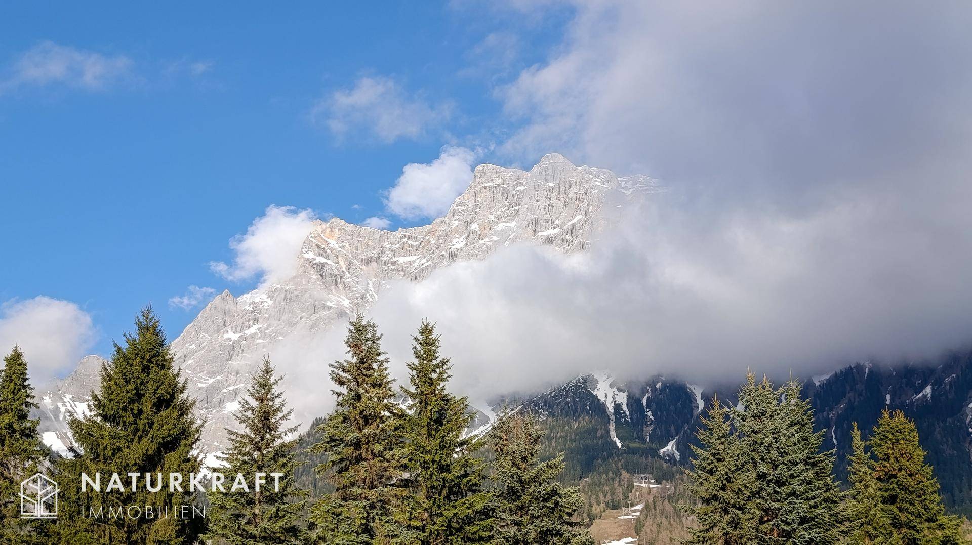 Majestätische schneebedeckte Berge, teilweise von Wolken verhüllt, mit Nadelbäumen im Vordergrund.
