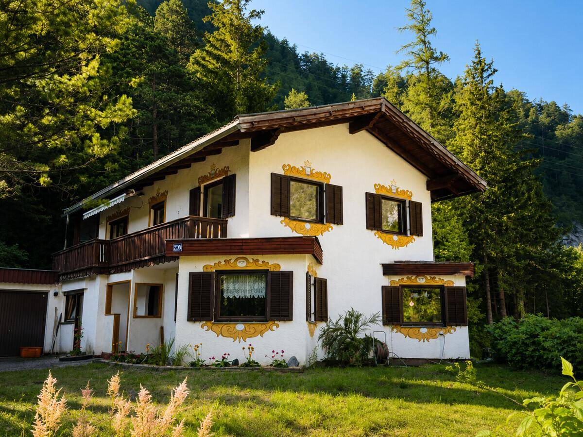 Traditionelles Haus mit weißer Fassade, dekorativen Malereien und Holzbalkon in alpiner Umgebung.