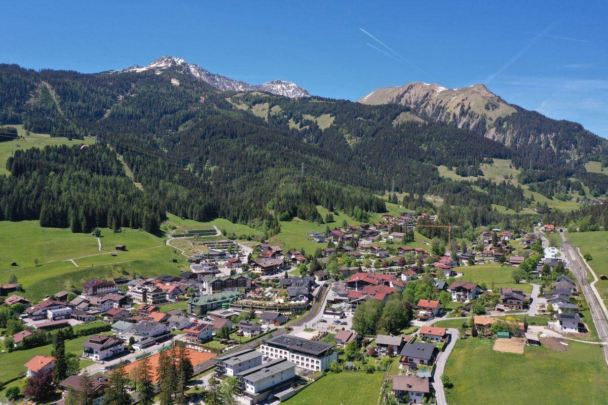 Panoramische Luftaufnahme einer Gemeinde in einem Tal, umgeben von bewaldeten Bergen und Wiesen.