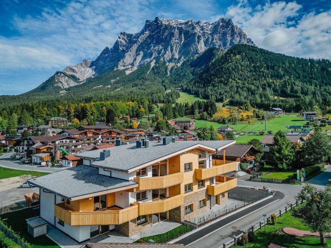 Luftaufnahme des Neubaus im alpinen Stil mit Holzverkleidung und Balkonen vor imposanter Bergkulisse.