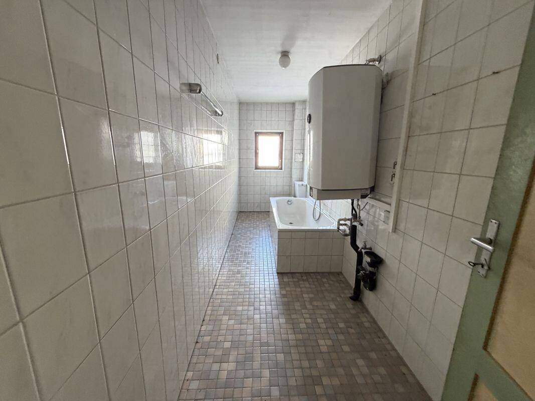 Langes Badezimmer mit weißen Wandfliesen, gemustertem Boden und einer Badewanne unter dem Fenster.