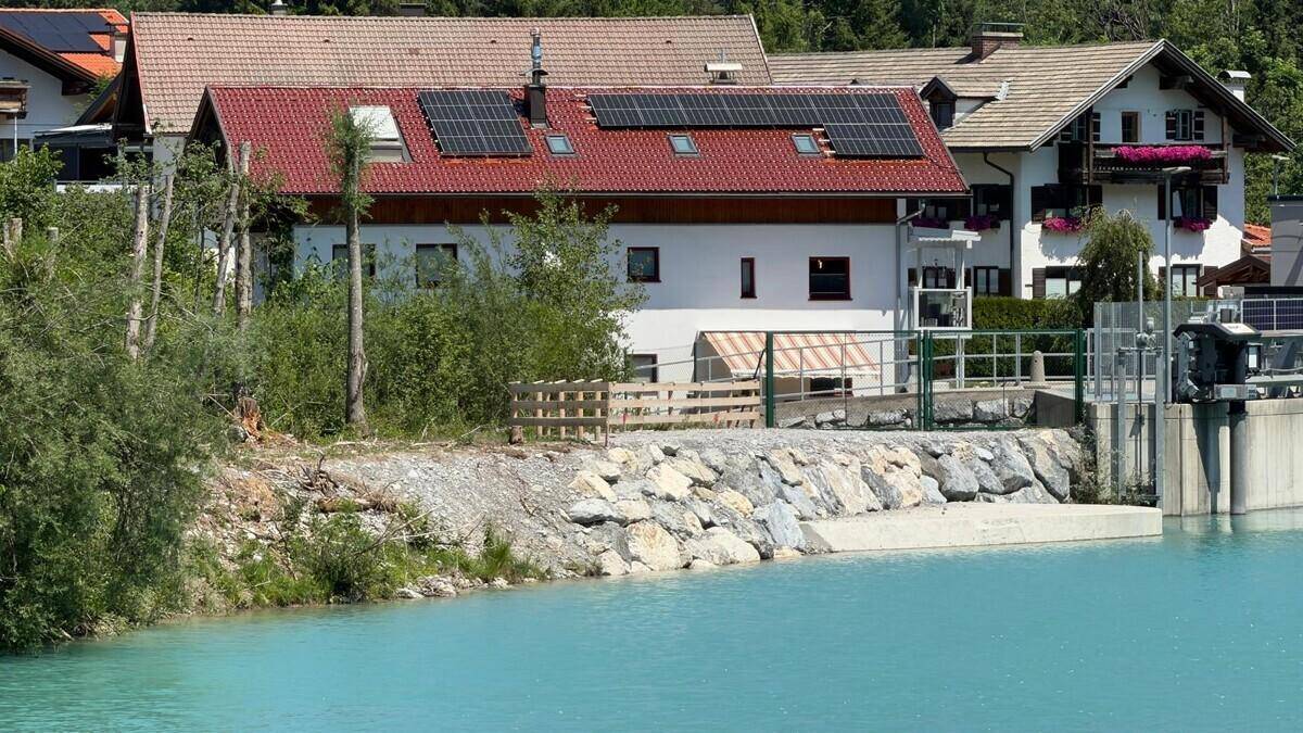Haus am Flussufer mit Solaranlage auf dem Dach und alpiner Bergkulisse im Hintergrund.