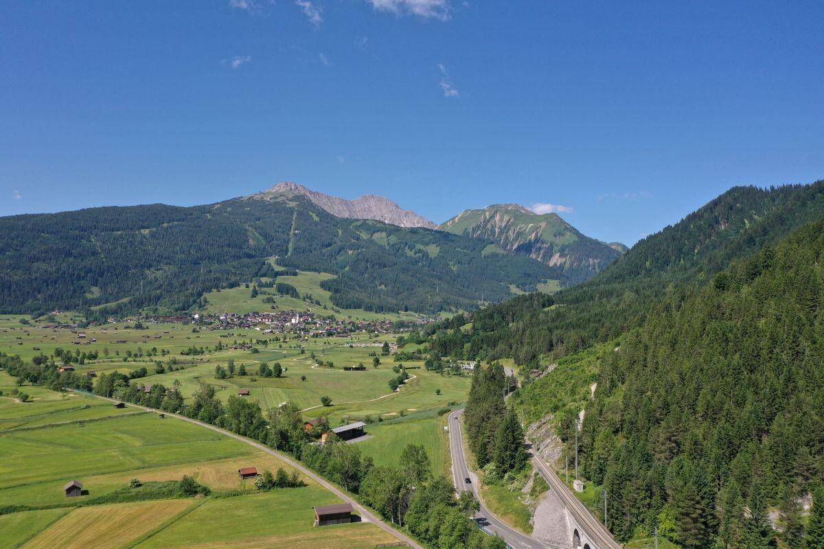 Luftaufnahme eines Tals mit Straße, Bahnlinie, Wäldern und einem Dorf vor einer Bergkulisse.