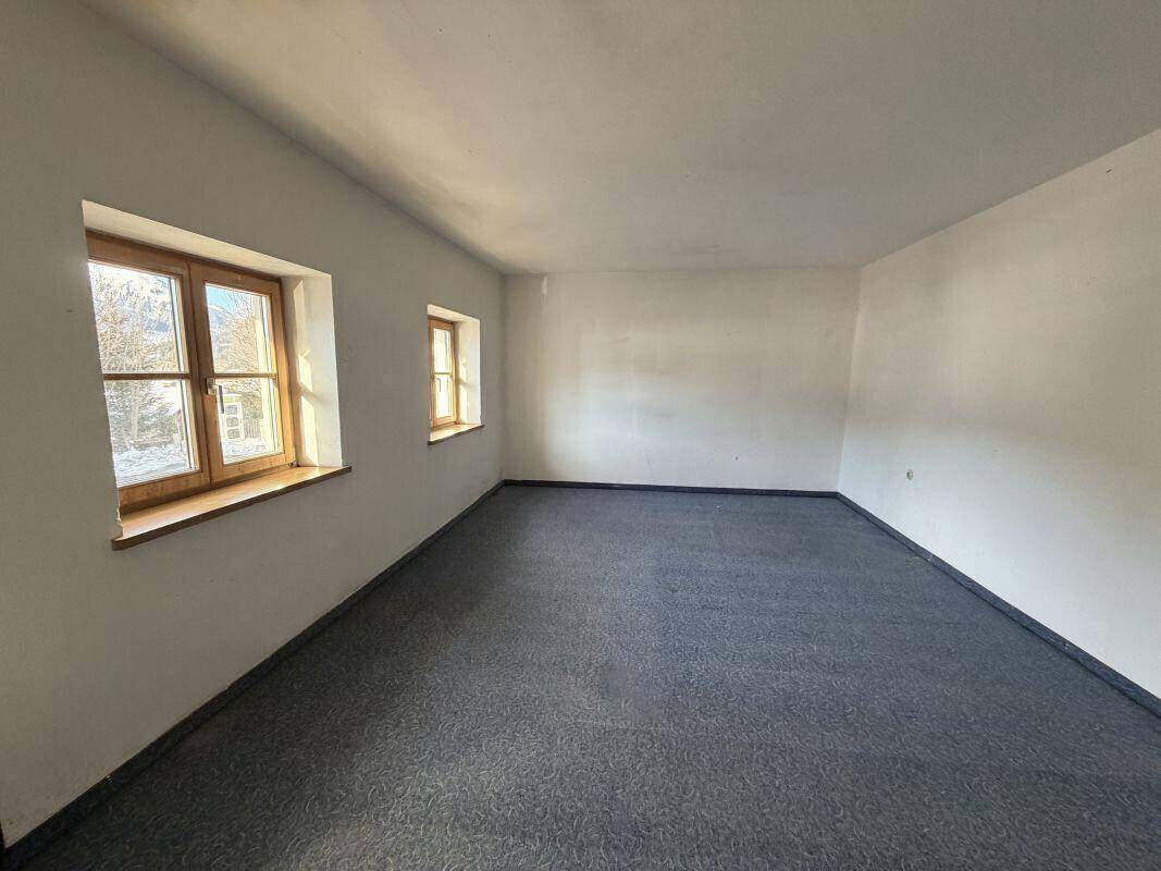 Leerer Raum mit dunklem Teppichboden, weißen Wänden und zwei Fenstern, die Tageslicht hereinlassen.