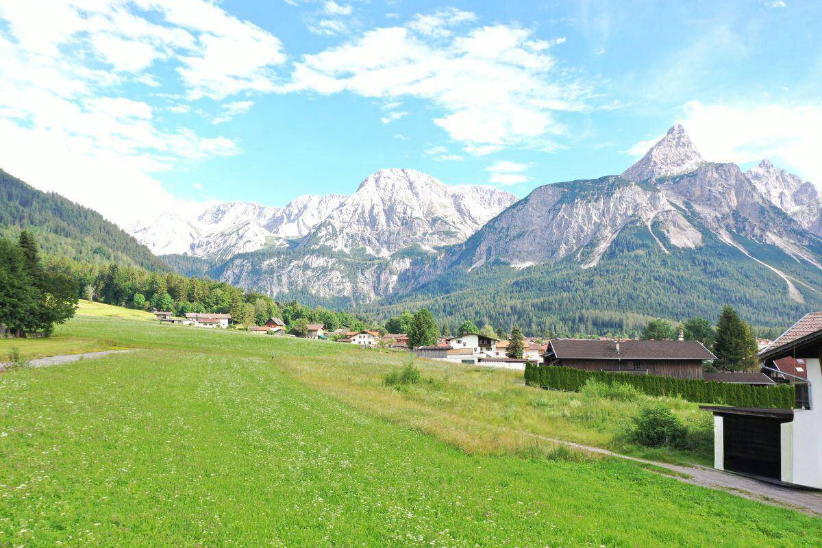 Weitläufige grüne Landschaft mit vereinzelten Häusern und majestätischen Bergen im Hintergrund unter blauem Himmel.