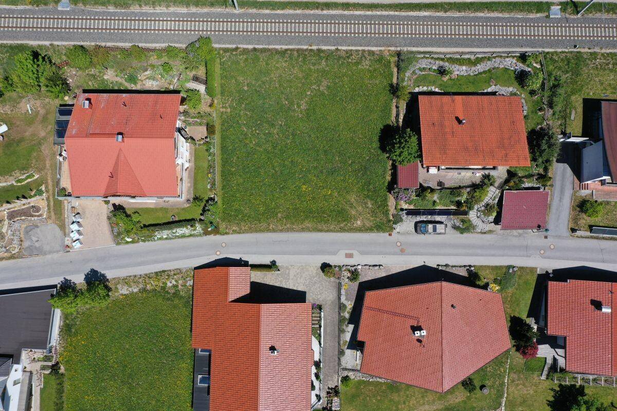 Luftaufnahme eines Wohngebiets mit Häusern, einer Straße, Bahngleisen und einem freien Grundstück.