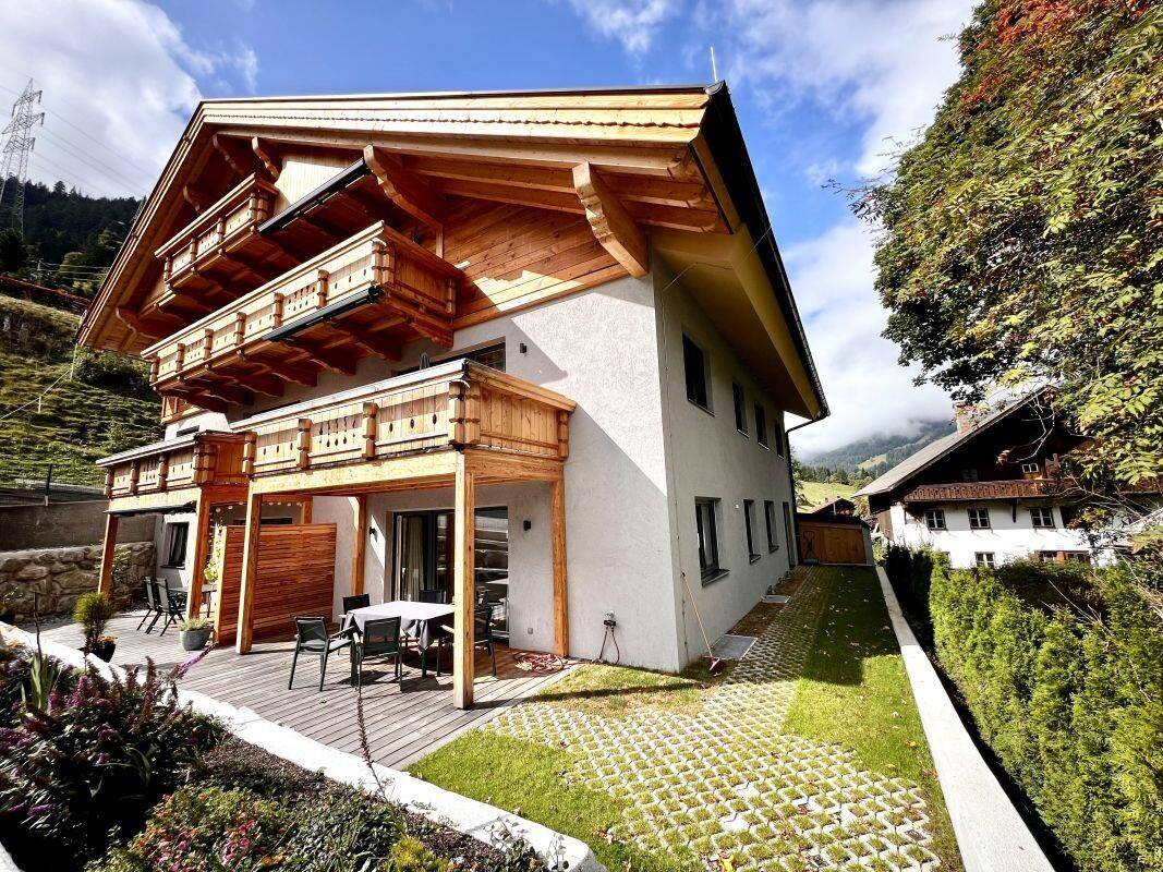 Modernes Gebäude mit traditionellen Holzbalkonen und einer Terrasse im Erdgeschoss, umgeben von alpiner Landschaft.