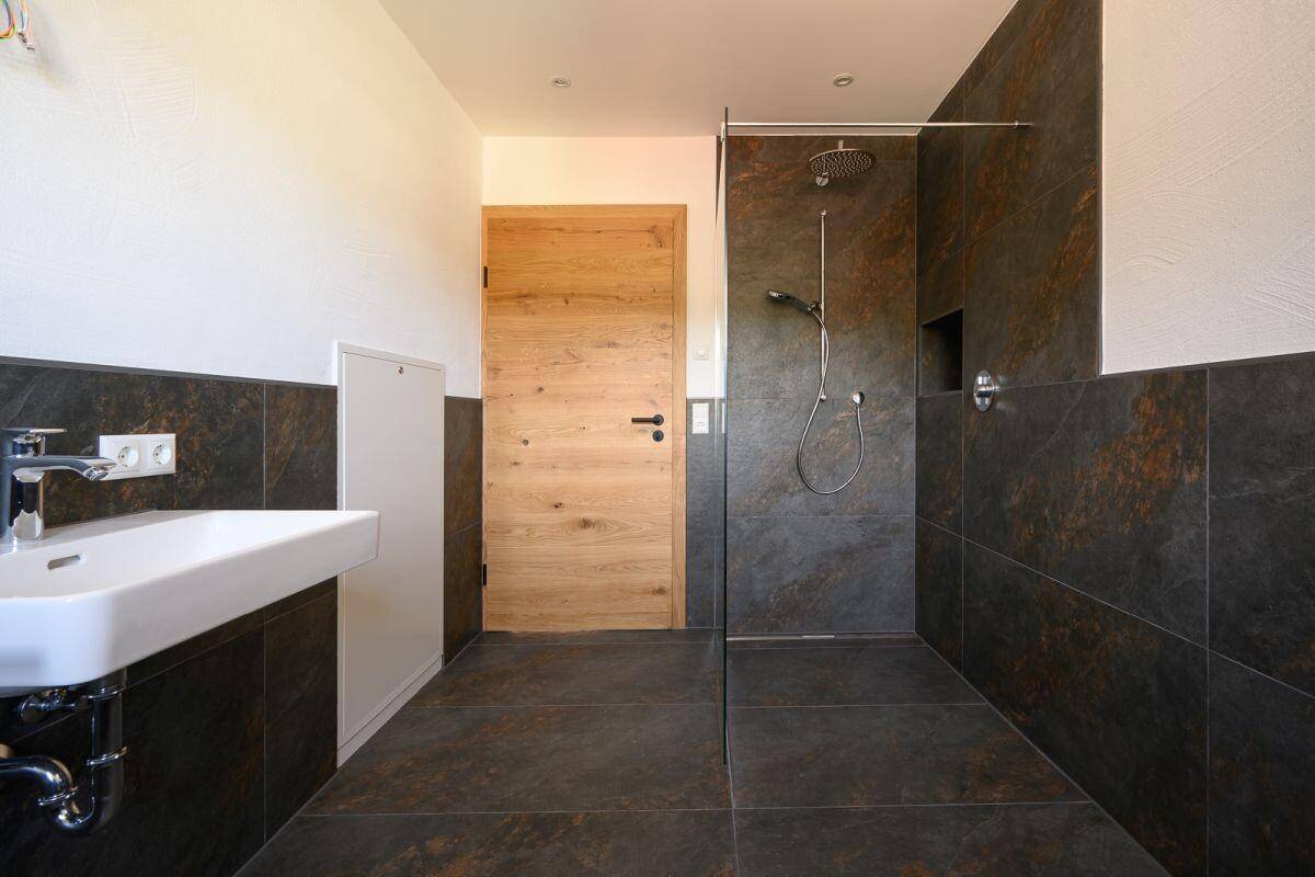 Elegantes Badezimmer mit dunklen Fliesen, moderner Dusche und Holztür.
