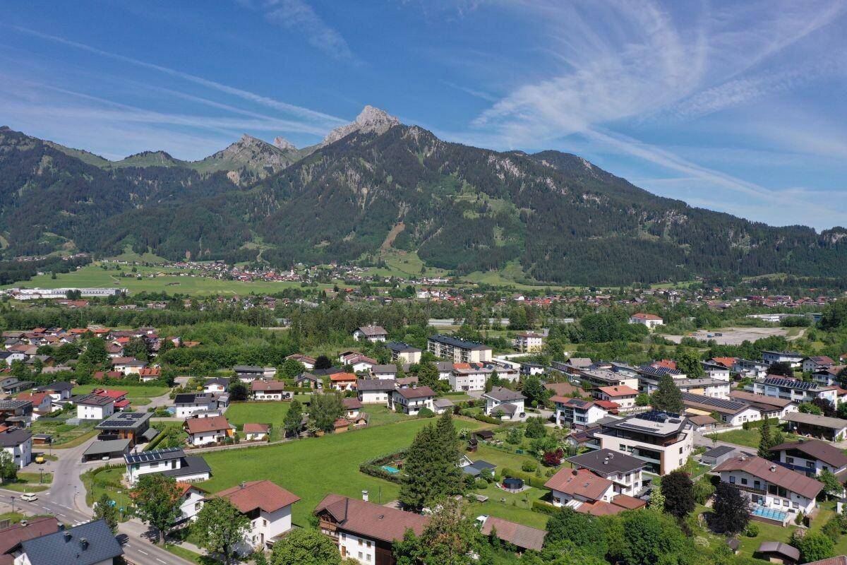 Weitläufige Luftaufnahme einer Siedlung mit vielfältigen Häusern vor der Kulisse imposanter Bergketten unter blauem Himmel.