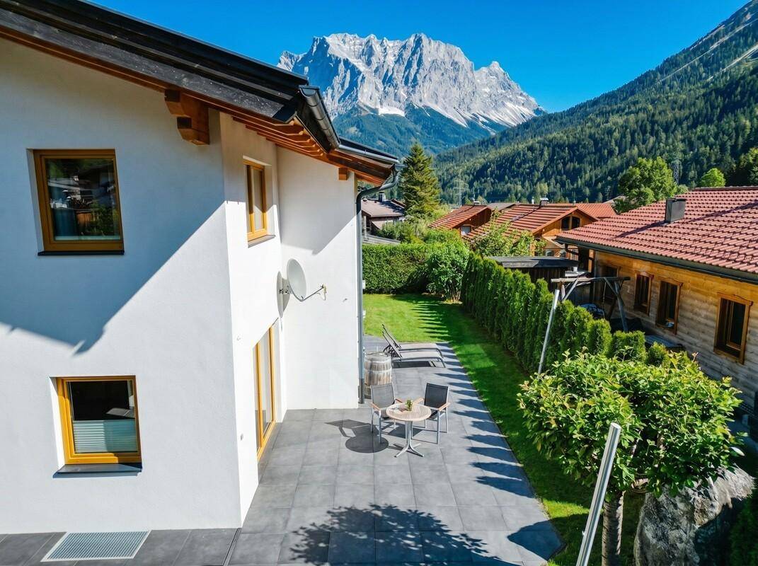 Hausansicht von der Seite mit gepflegtem Garten, Terrasse und majestätischem Bergpanorama.
