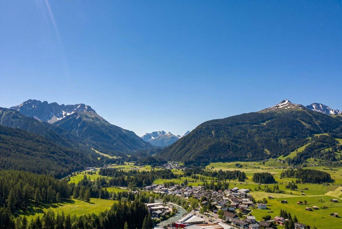 Weitläufige Luftaufnahme eines Tals mit einer Siedlung, umgeben von Bergen und Wäldern.