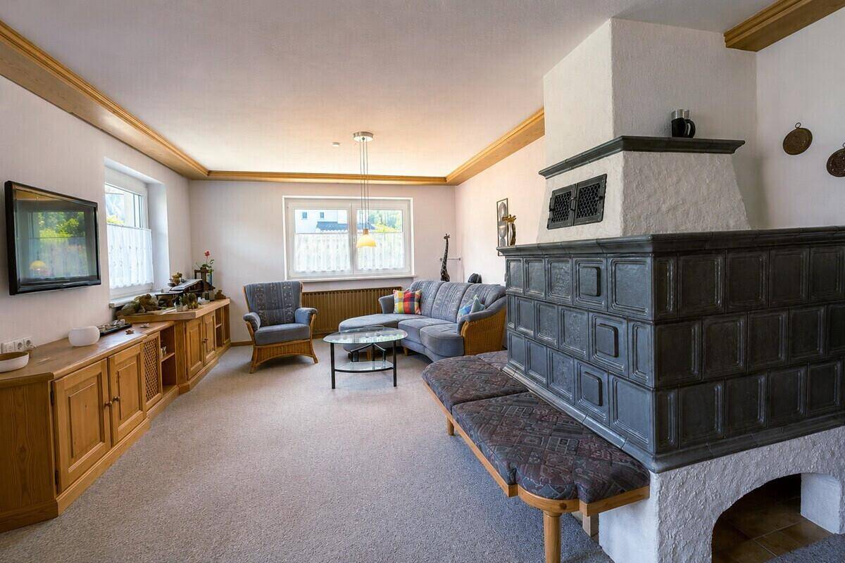Geräumiges Wohnzimmer mit Kachelofen, Teppichboden und traditionellen Holzmöbeln.