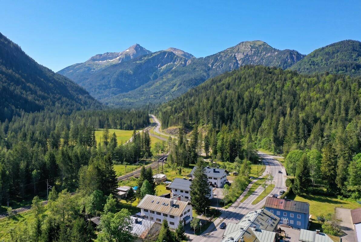 Malerisches Tal mit Straße, Fluss, Häusern und majestätischen Bergen unter blauem Himmel.