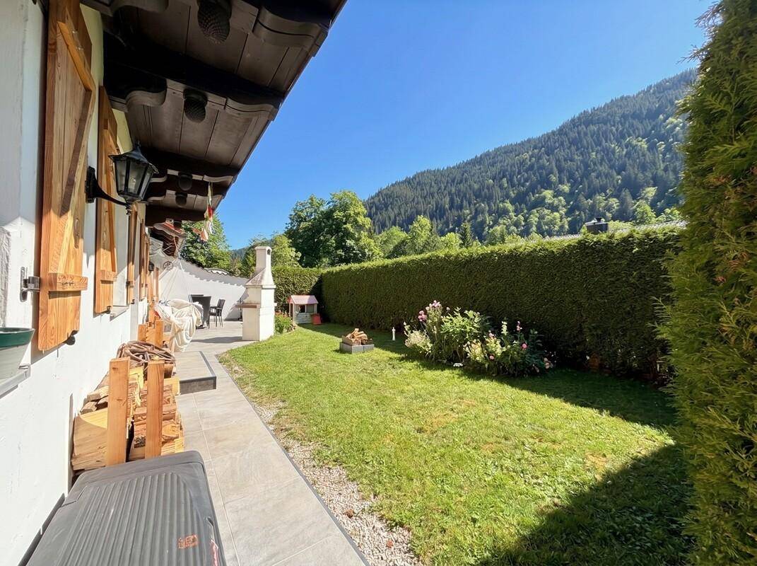 Seitlicher Gartenbereich mit Rasenfläche, gepflasterter Terrasse und Bergblick, umgeben von Hecken.