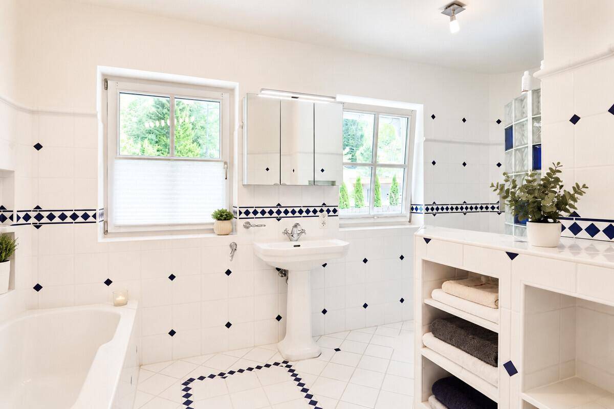 Helles Badezimmer mit weiß-blauen Fliesen, Standwaschbecken, Badewanne und zwei Fenstern.