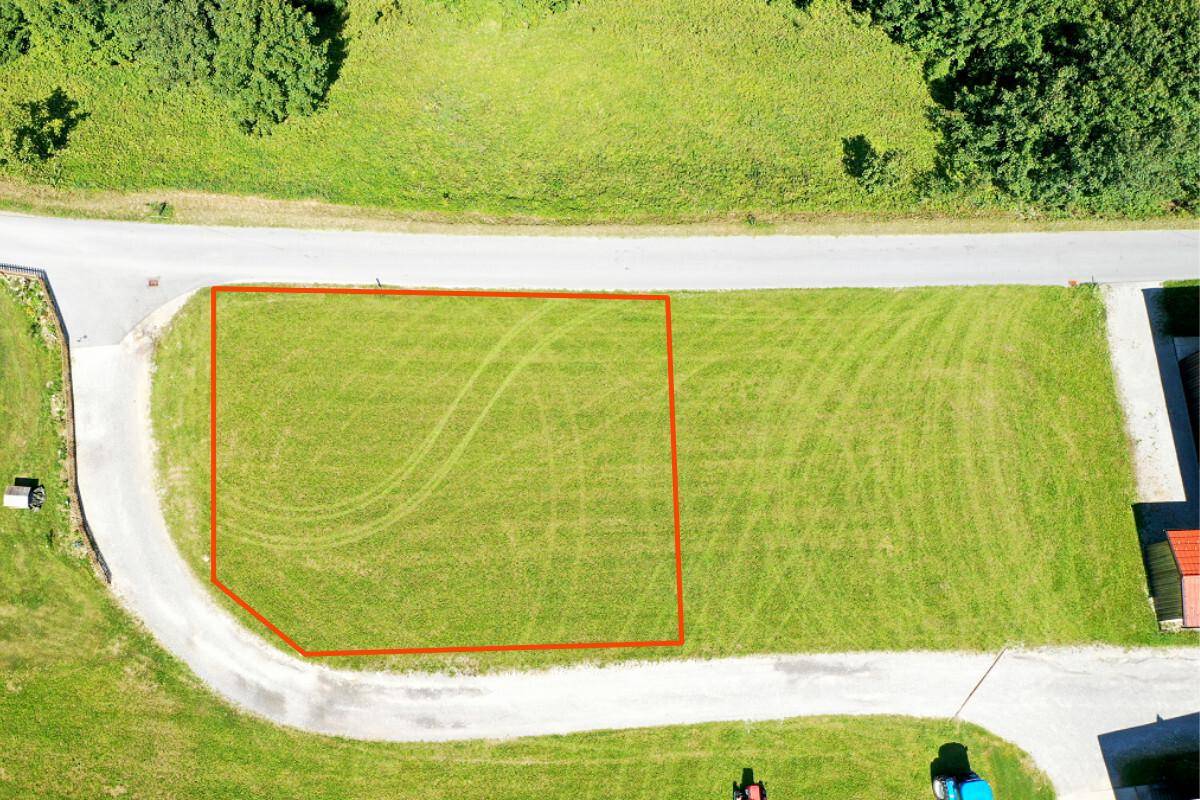 Luftaufnahme eines grünen Baugrundstücks, rot umrandet, mit Zufahrtsstraße.