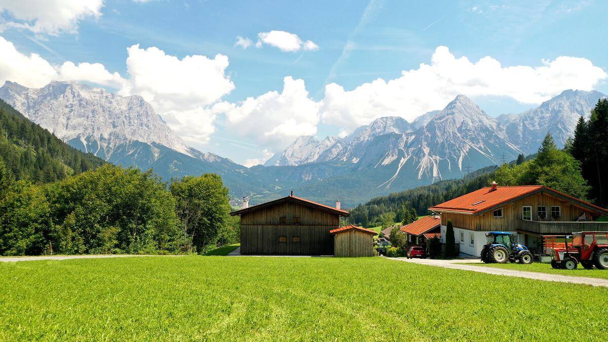 Ländliche Szene mit traditionellen Häusern, grünen Wiesen und landwirtschaftlichen Fahrzeugen vor Bergkulisse.