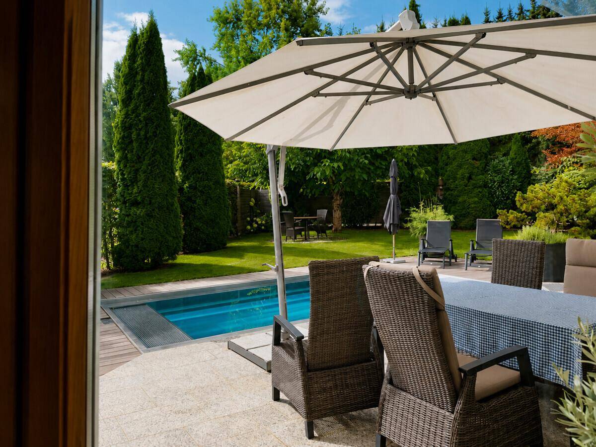 Blick vom Innenbereich auf den grünen Garten mit Pool, Terrasse und hohen Bäumen.