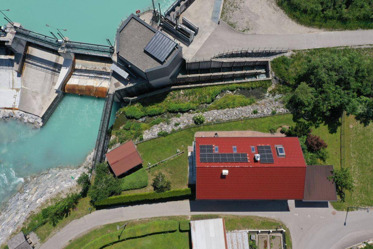 Luftaufnahme eines Hauses mit rotem Dach und Solaranlage, direkt am Fluss und Staudamm gelegen.