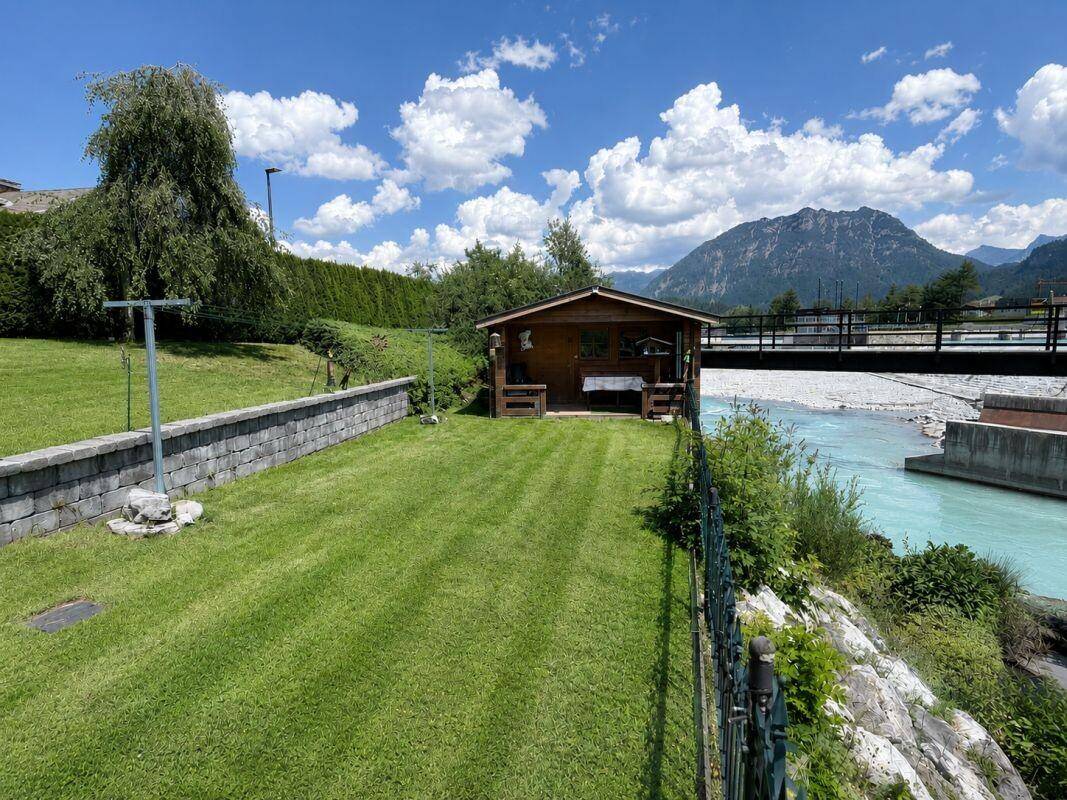 Großer, gepflegter Garten mit Rasenfläche, Gartenhaus und Flussblick vor Bergkulisse.