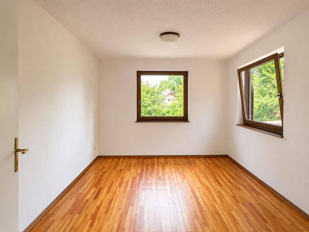 Freundliches Zimmer mit Holzparkettboden und zwei Fenstern, die viel Tageslicht hereinlassen.
