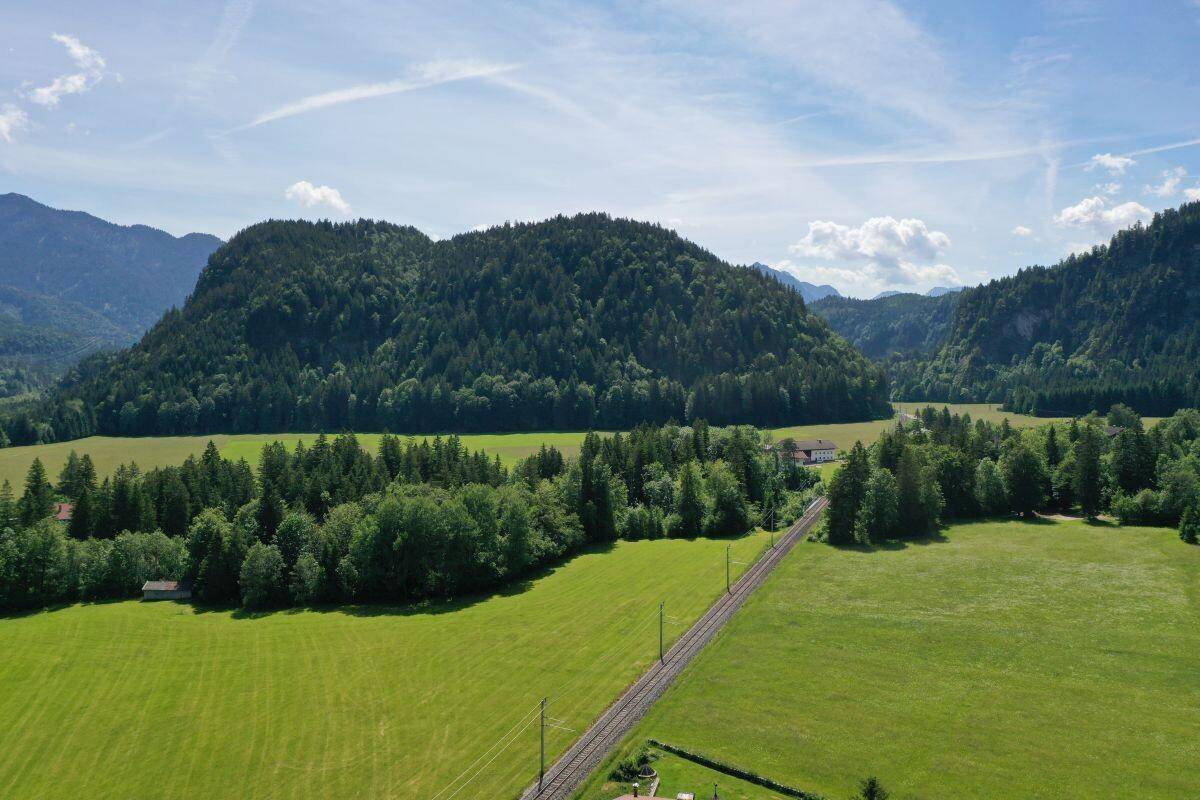 Weitläufige Landschaft mit grünen Feldern, dichten Wäldern und einer Bahnlinie vor einer Bergkulisse.