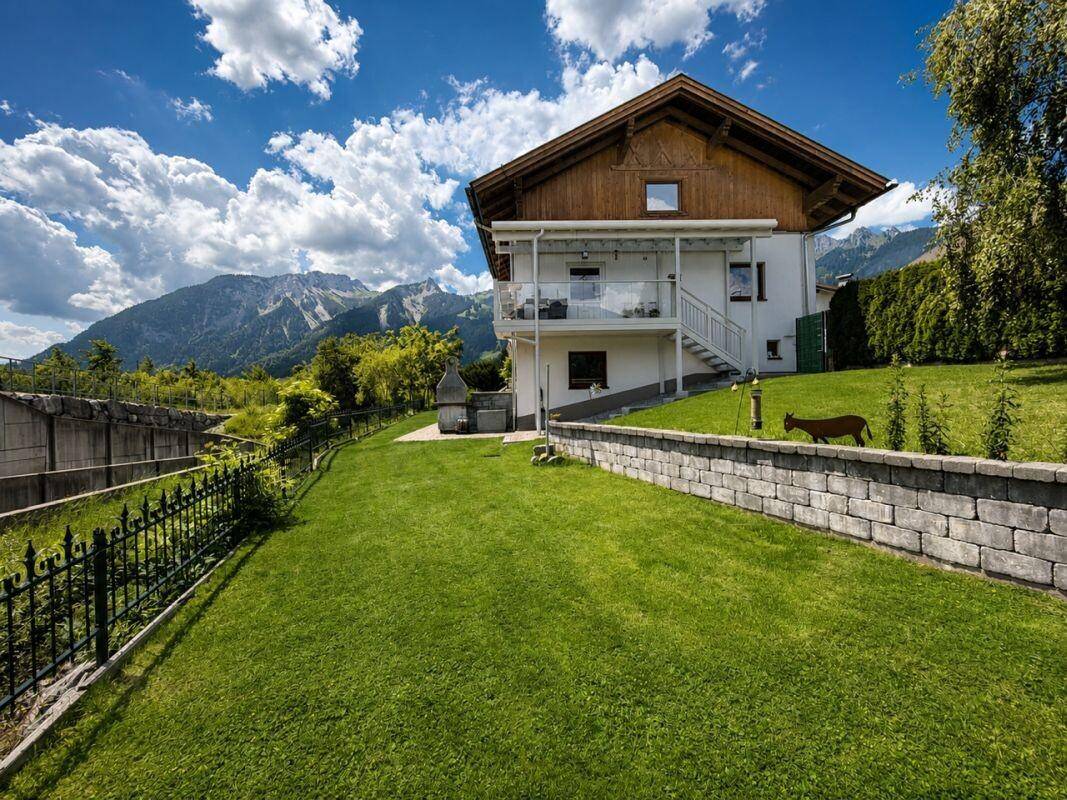 Hinteransicht des Hauses mit großem Garten, Balkon und Bergblick unter blauem Himmel.