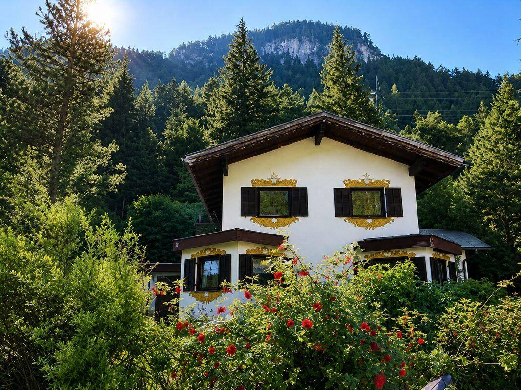 Alpines Haus mit traditioneller Architektur, umgeben von üppiger Vegetation und Bergen im Hintergrund.